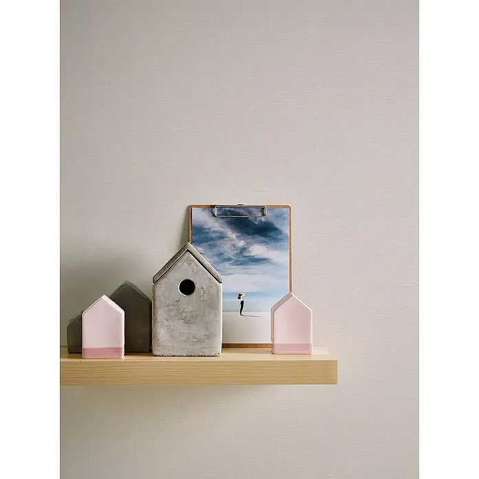 A.S. Création Casual Living Vliestapete Textilstruktur - Cremeweiß, Uni, 10,05 x 0,53 m - Stillleben mit Deko-Häusern und einem Foto auf einem Holzregal vor einer schlichten Wand.