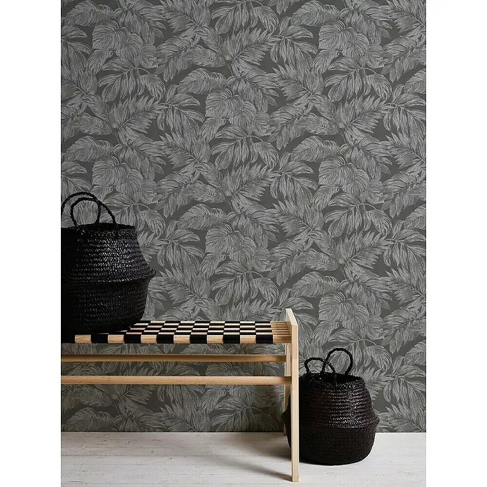 A.S. Création Casual Living Vliestapete - Anthrazit/Grau/Silber, Floral, 10,05 x 0,53 m - Eine stilvolle Szene mit zwei schwarzen Körben und einer hellen Holzbank vor einer grauen Blättertapete, die eine moderne Ästhetik vermittelt.