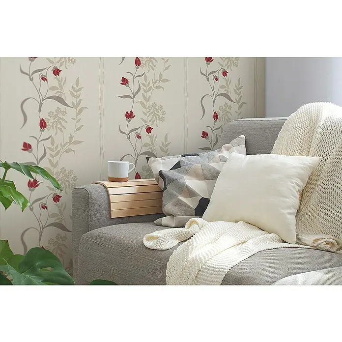 A.S. Création Casual Living Vliestapete - Creme/Rot/Beige, Floral, 10,05 x 0,53 m - Eine Nahaufnahme eines grauen Sofas mit einer weißen Strickdecke, geometrischen Kissen und einem Beistelltisch mit einer Kaffeetasse, der ein gemütliches Ambiente erzeugt.