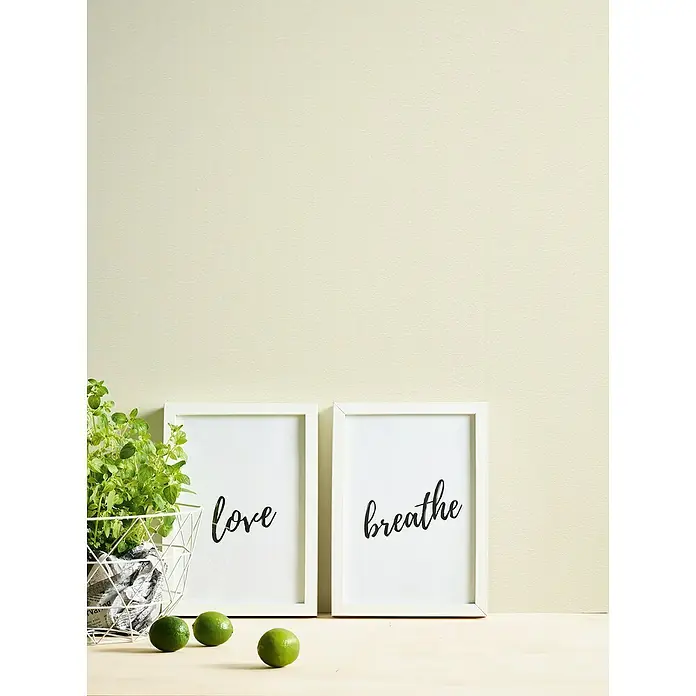 A.S. Création Casual Living Vliestapete - Beigeweiß, Uni, 10,05 x 0,53 m - Nahaufnahme von zwei gerahmten Kunstdrucken mit den Aufschriften "Love" und "Breathe", umgeben von einer Pflanze und Limetten.
