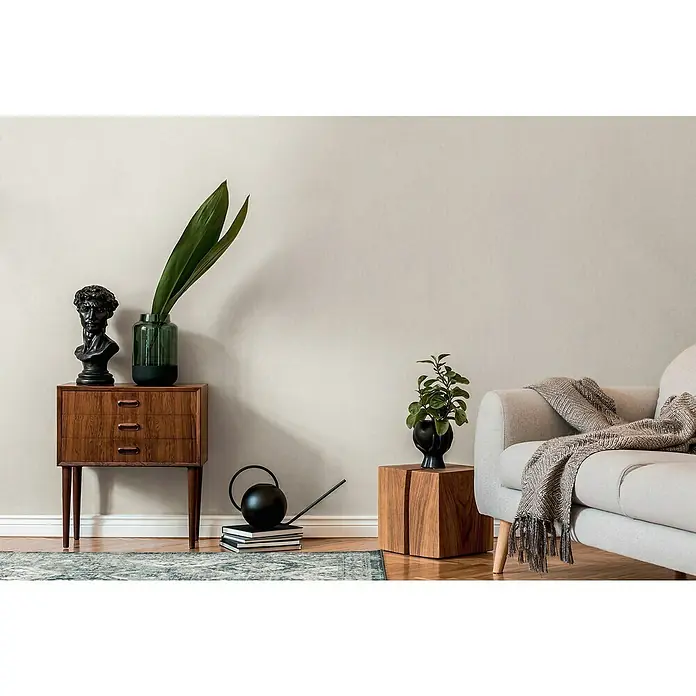 A.S. Création Casual Living Vliestapete - Cremiges Weiß, Uni, 10,05 x 0,53 m - Einrichtungsszene mit einem braunen Holzschrank, einer Skulptur, einer Pflanze und einem beigen Sofa mit einer dekorativen Decke.