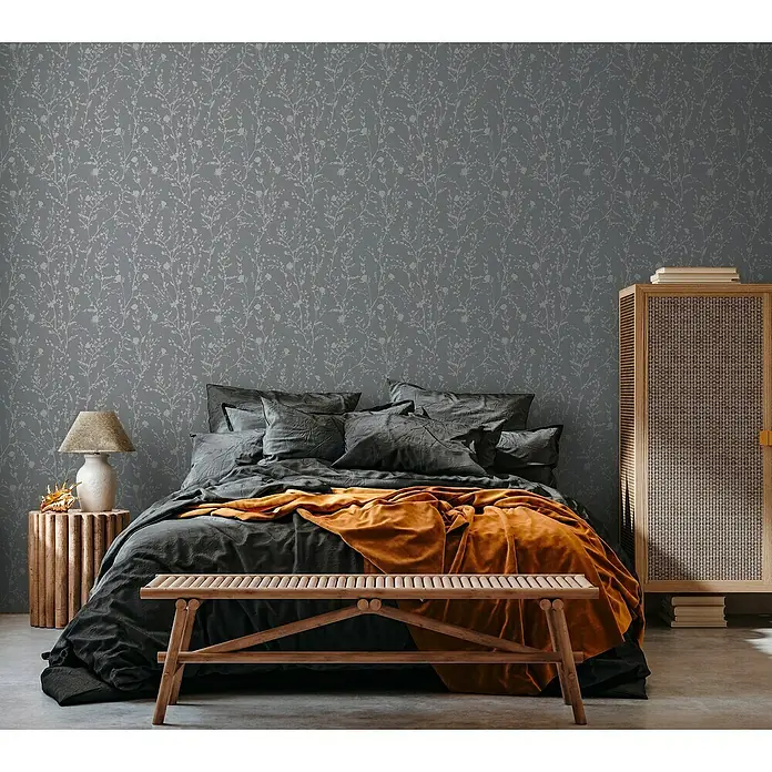 A.S. Création Casual Living Vliestapete - Silbergrau, Floral, 10,05 x 0,53 m - Schlafzimmer mit grauer Blumentapete, dunkler Bettwäsche und einer orangefarbenen Decke für ein stilvolles und gemütliches Ambiente.