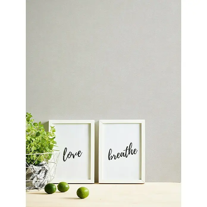 A.S. Création Casual Living Vliestapete - Grau/Hellgrau, Uni, 10,05 x 0,53 m - Dekorative "Love, Breathe" gerahmte Wandkunst neben einer Korbpflanze und Limetten, die eine ruhige und stilvolle Atmosphäre ausstrahlen.