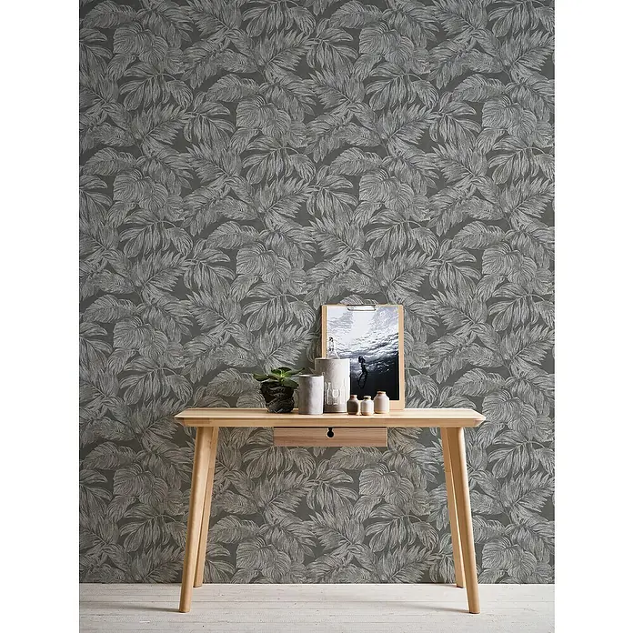 A.S. Création Casual Living Vliestapete - Anthrazit/Grau/Silber, Floral, 10,05 x 0,53 m - Nahaufnahme eines hellen Holzschreibtisches mit Schublade vor einer Wand mit Blättermuster.