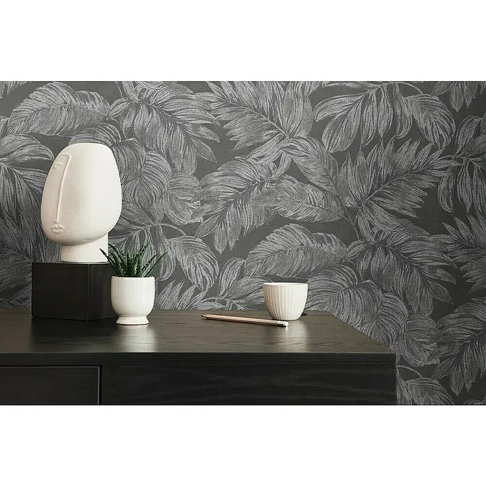 A.S. Création Casual Living Vliestapete - Anthrazit/Grau/Silber, Floral, 10,05 x 0,53 m - Nahaufnahme einer minimalistischen Raumdekoration mit einer weißen Skulptur und Zimmerpflanzen auf einer dunklen Kommode vor floraler Tapete.