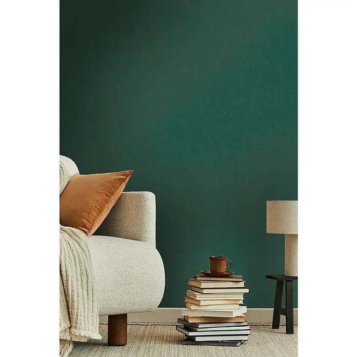 A.S. Création Casual Living Vliestapete - Waldgrün, Uni, 10,05 x 0,53 m - Nahaufnahme eines stilvollen Wohnzimmers mit einem Bücherstapel, einer Lampe und einem bequemen Sessel.