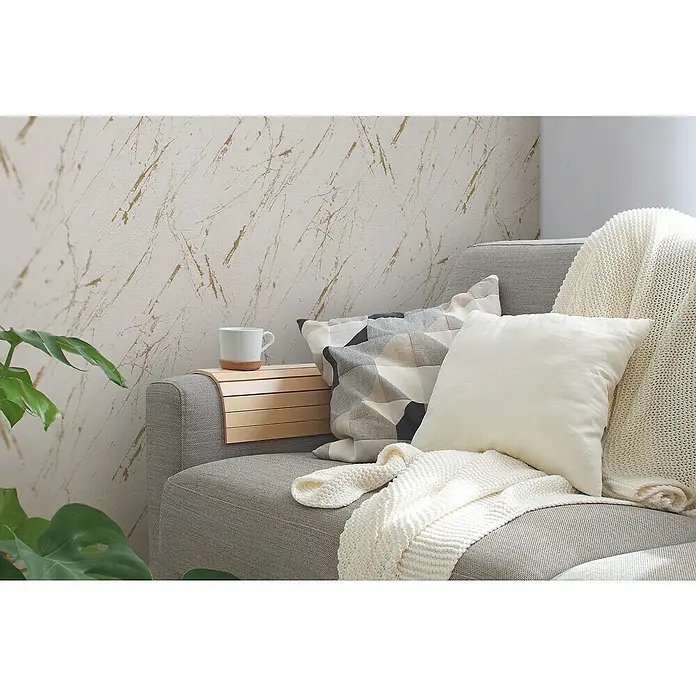 A.S. Création Casual Living Vliestapete Marmor - Beige/Gold/Metallic, Steinoptik, 10,05 x 0,53 m - Eine gemütliche Szene mit einem grauen Sofa, dekoriert mit Kissen und einer weichen Decke, sowie einem Beistelltischchen mit einer Tasse.