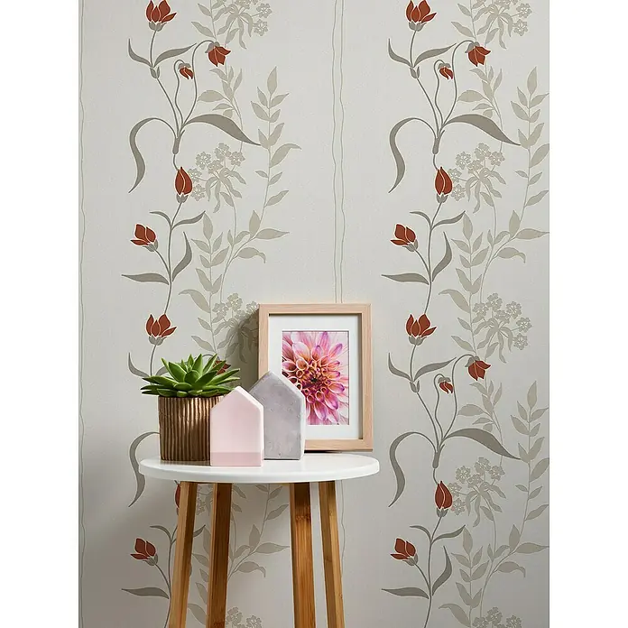 A.S. Création Casual Living Vliestapete - Creme/Rot/Beige, Floral, 10,05 x 0,53 m - Nahaufnahme eines dekorierten, runden Beistelltisches mit Topfpflanze, Deko-Häusern und Bilderrahmen auf einem gemusterten Hintergrund.