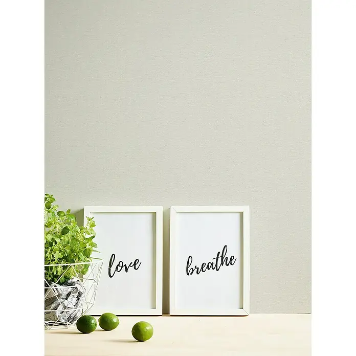A.S. Création Casual Living Vliestapete - Cremiges Weiß, Uni, 10,05 x 0,53 m - Deko-Arrangement mit "Love" und "Breathe" Kunstdrucken, Limetten und einer Pflanze in einem Drahtkorb auf einem Tisch.