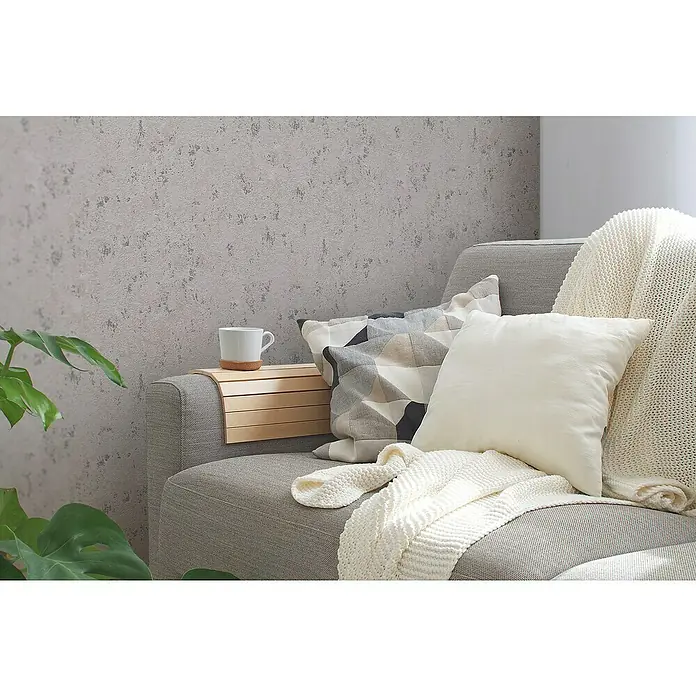 A.S. Création Casual Living Vliestapete Struktur - Grau/Beige/Silber, Uni, 10,05 x 0,53 m - Nahaufnahme eines grauen Sofas mit einem weißen Kissen, einer Decke und einem Beistelltisch mit einer Tasse darauf vor einer strukturierten Wand.