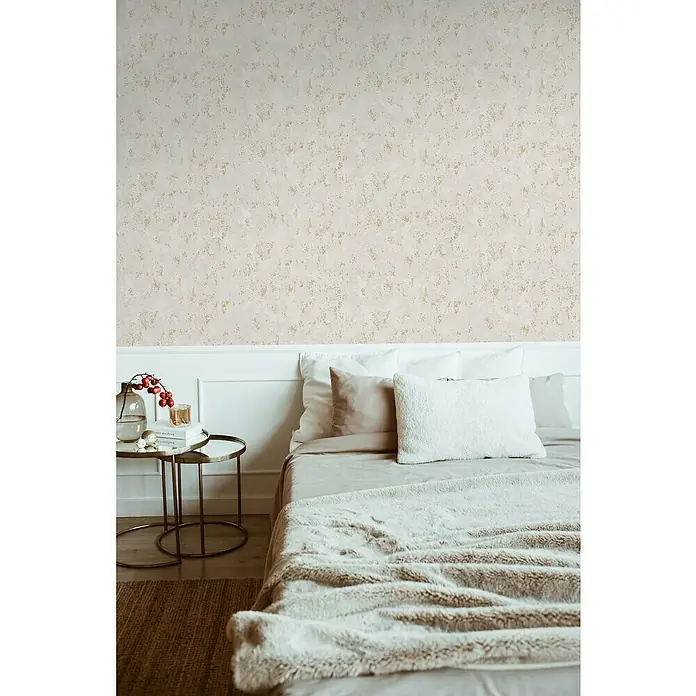 A.S. Création Casual Living Vliestapete Struktur - Grau/Gold/Metallic, Uni, 10,05 x 0,53 m - Nahaufnahme eines Bettes mit beigen Kissen, einer weichen Decke und einem Beistelltisch mit Dekoration vor einer strukturierten Wand.