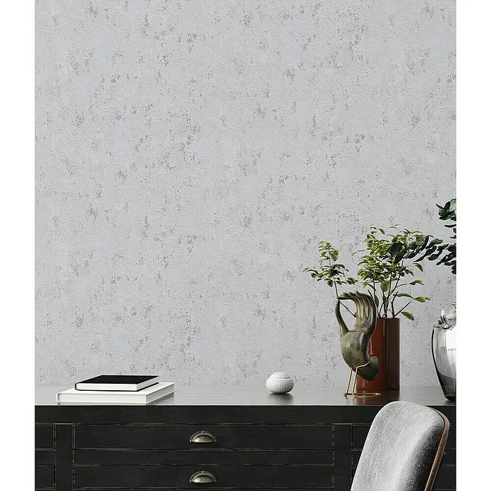 A.S. Création Casual Living Vliestapete Struktur - Grau/Silber/Metallic, Uni, 10,05 x 0,53 m - Nahaufnahme einer dunklen Kommode mit dekorativen Accessoires wie Pflanzen, Büchern und einer Skulptur vor einer strukturierten, hellgrauen Wand.