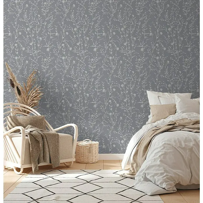 A.S. Création Casual Living Vliestapete - Silbergrau, Floral, 10,05 x 0,53 m - Ein Schlafzimmer mit einer grauen Wandtapete mit weißen Blütenmotiven. Das Zimmer ist mit einem Bett und einem Sessel ausgestattet.