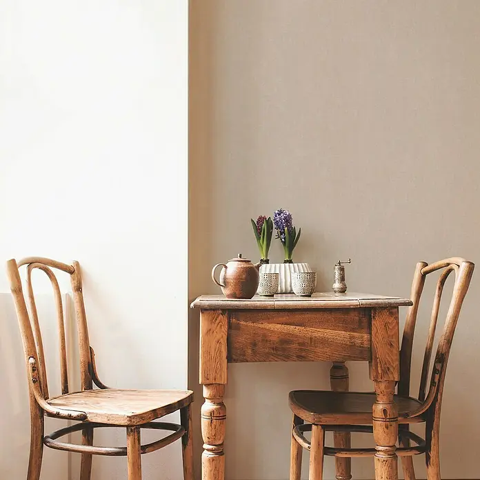 A.S. Création Casual Living Vliestapete - Harmonisches Beige, Uni, 10,05 x 0,53 m - Ein Vintage-Esstisch aus Holz mit Teekanne, Gewürzmühle und zwei Blumenvasen, umgeben von zwei Holzstühlen.