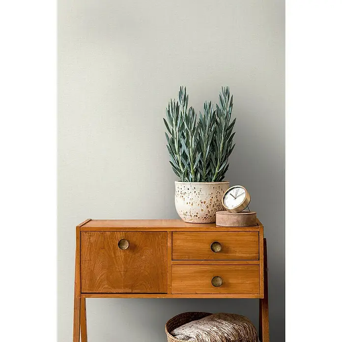 A.S. Création Casual Living Vliestapete - Cremiges Weiß, Uni, 10,05 x 0,53 m - Ein stilvolles Sideboard aus Holz mit einer Topfpflanze und einer Uhr darauf, die das warme Ambiente des Raumes betonen.