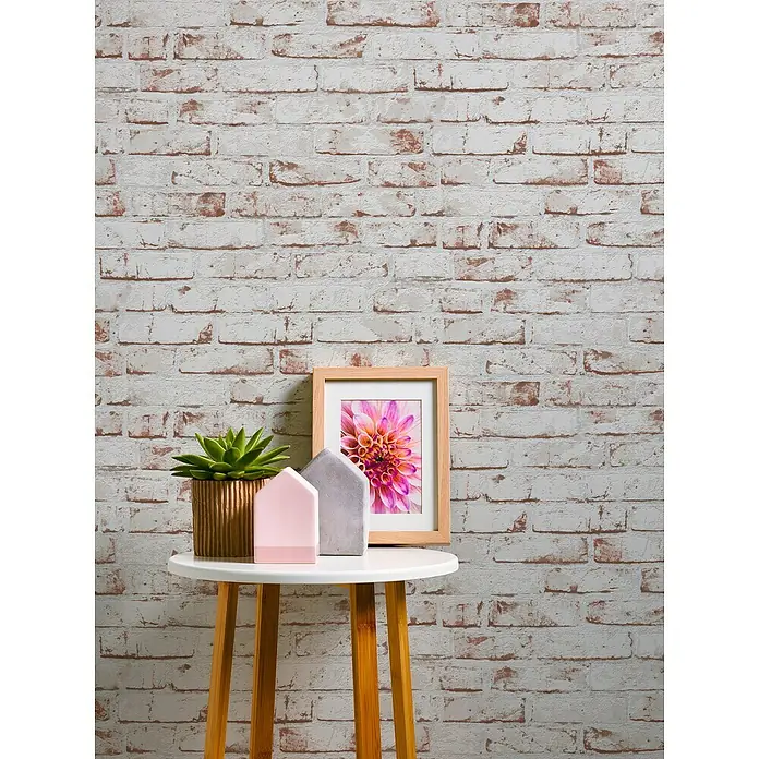 A.S. Création Casual Living Vliestapete New England Klinker - Weiß/Rot, Steinoptik, 10,05 x 0,53 m - Dekoratives Stillleben mit einem Bilderrahmen mit Blumenmotiv, einer Sukkulente und zwei Zierhäusern auf einem Beistelltisch vor einer Backsteinwand.