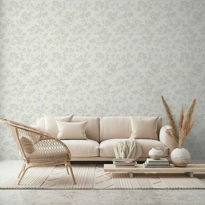 A.S. Création Casual Living Vliestapete Blümchen - Weiß/Grün/Grau, Floral, 10,05 x 0,53 m - Wohnzimmer mit einem beigen Sofa, einem Rattan-Stuhl und einem Couchtisch mit Dekor, vor einer Wand mit floralem Muster.