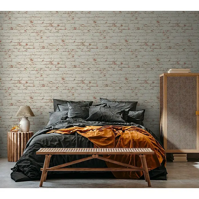 A.S. Création Casual Living Vliestapete New England Klinker - Weiß/Rot, Steinoptik, 10,05 x 0,53 m - Schlafzimmer mit Ziegelwand, Bett mit grauer Bettwäsche und orangefarbener Decke sowie einer Holzbank. Schrank mit Bücherstapel.