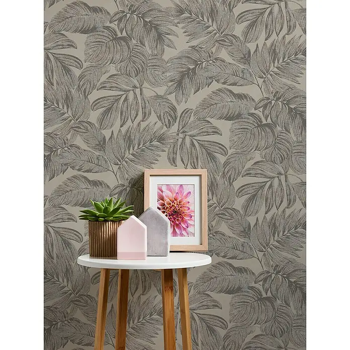 A.S. Création Casual Living Vliestapete - Braun/Grau/Beige, Floral, 10,05 x 0,53 m - Nahaufnahme eines eleganten Beistelltisches mit Dekorationen wie einer Sukkulente, Bilderrahmen und Zierhäusern vor einer Blättertapete.