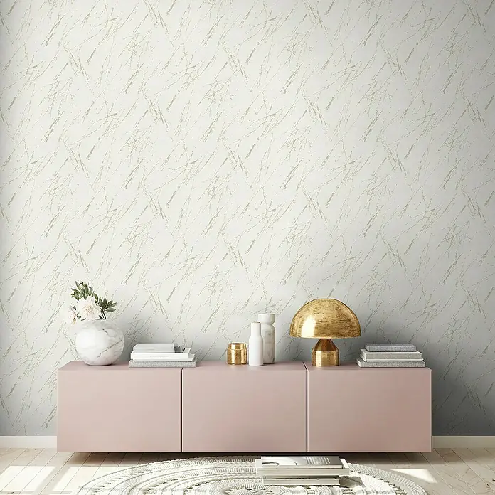 A.S. Création Casual Living Vliestapete Marmor - Weiß/Champagner/Metallic, Steinoptik, 10,05 x 0,53 m - Nahaufnahme eines rosa Sideboards mit Marmorvase, Lampe und Büchern vor einer strukturierten Tapete.