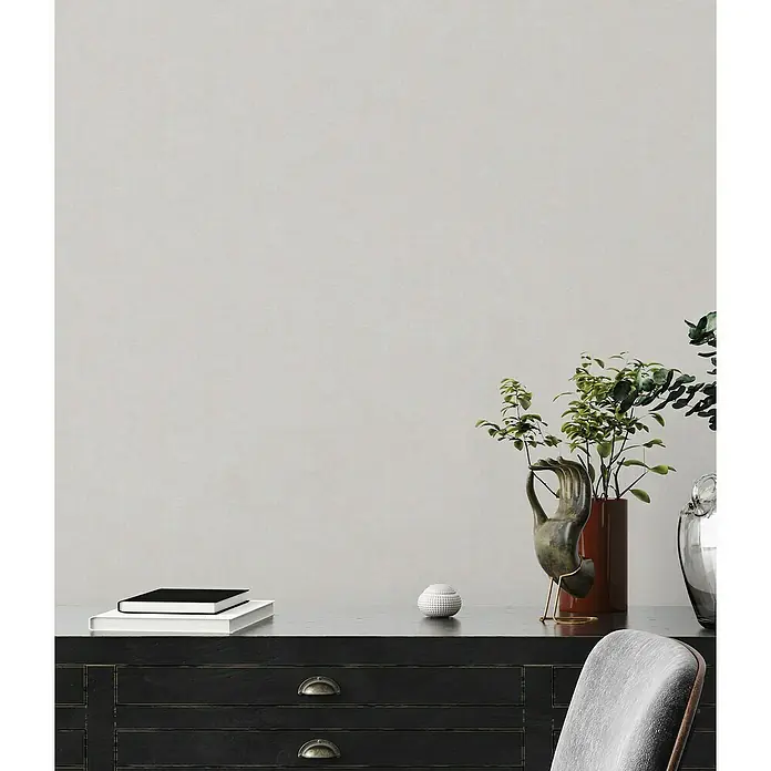 A.S. Création Casual Living Vliestapete - Grau/Hellgrau, Uni, 10,05 x 0,53 m - Stillleben mit Dekorationen auf einem schwarzen Schrank, darunter eine Skulptur und Grünpflanzen in einer Vase.