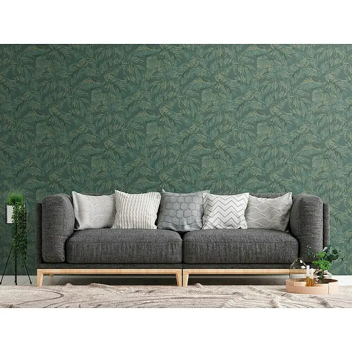 A.S. Création Casual Living Vliestapete - Grün/Gold, Floral, 10,05 x 0,53 m - Graues Sofa mit Kissen vor einer Wand mit Blättermuster, das eine stilvolle und gemütliche Wohnatmosphäre schafft.