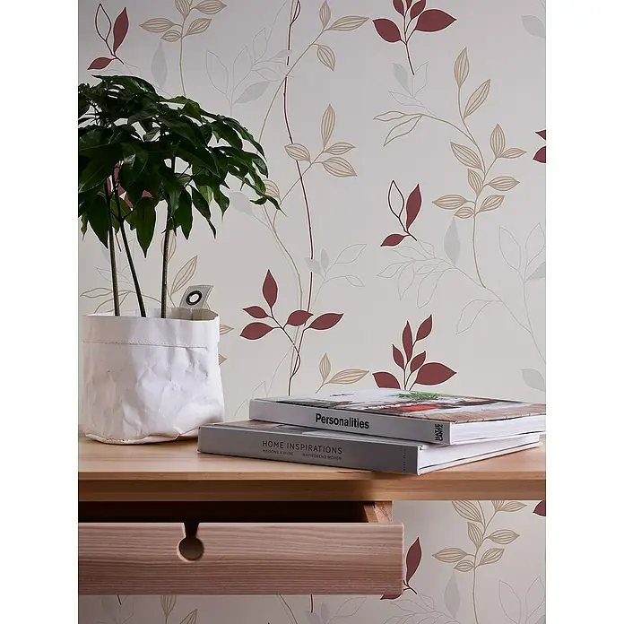 A.S. Création Casual Living Vliestapete Blätter - Creme/Rot/Beige, Floral, 10,05 x 0,53 m - Nahaufnahme eines Tisches mit einer Zimmerpflanze, zwei Büchern ("Personalities", "Home Inspirations") und einer Schublade vor einer floralen Tapete.