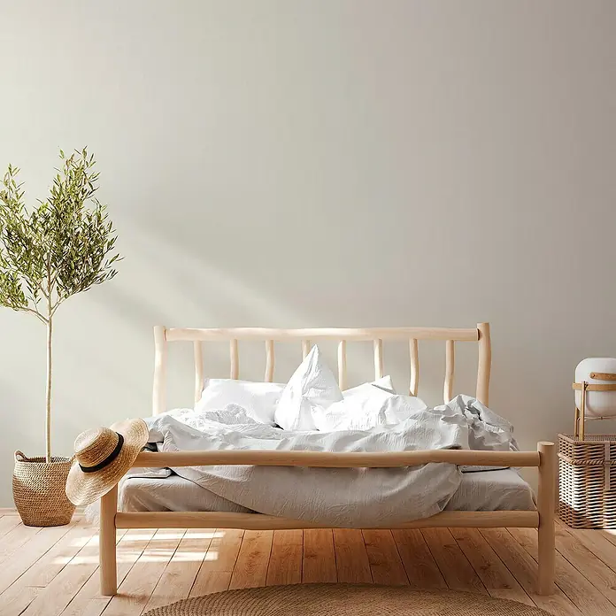 A.S. Création Casual Living Vliestapete - Cremiges Weiß, Uni, 10,05 x 0,53 m - Elegantes Schlafzimmer mit einem hellen Holzrahmenbett, das mit weißer Bettwäsche und Kissen einladend wirkt. Eine grüne Pflanze und ein Strohhut ergänzen das Ambiente.