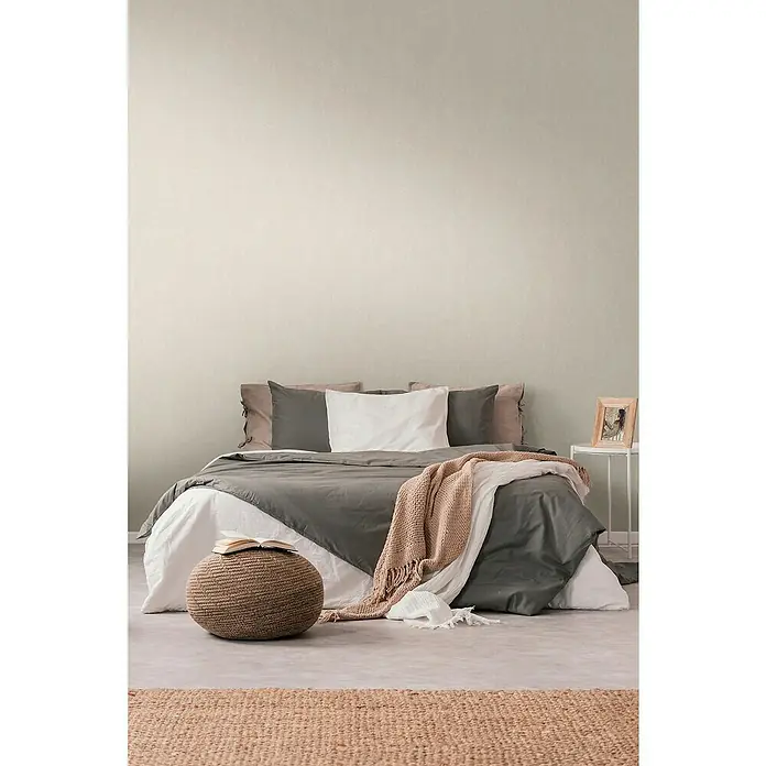 A.S. Création Casual Living Vliestapete - Harmonisches Beige, Uni, 10,05 x 0,53 m - Schlafzimmer mit einem grauen Tagesdecke-Set, einem beigen Hocker und einem hellbraunen Teppich für eine gemütliche Atmosphäre.