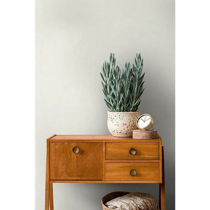 A.S. Création Casual Living Vliestapete - Freundliches Weiß, Uni, 10,05 x 0,53 m - Holzkommode mit einem einzelnen Schrank, zwei Schubladen, einer Topfpflanze, einer Uhr und einem Korb mit einer Decke.