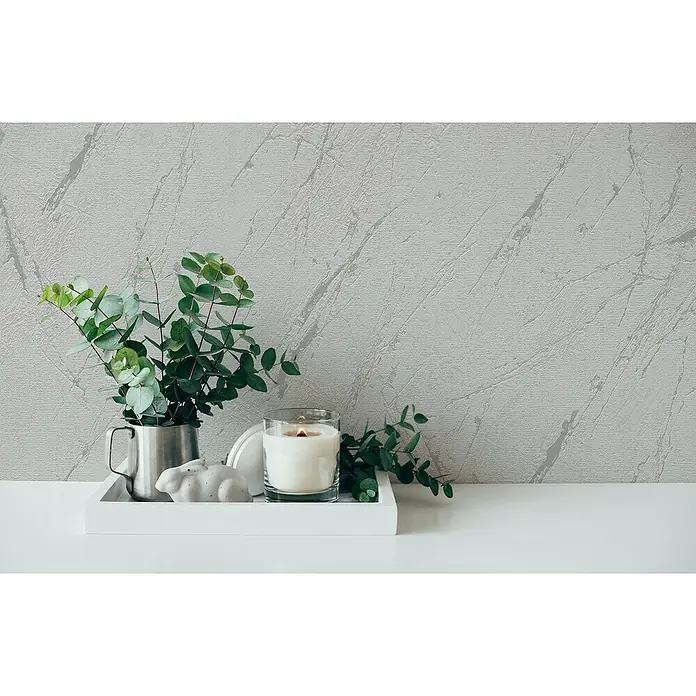 A.S. Création Casual Living Vliestapete Marmor - Silbergrau-Metallic, Steinoptik, 10,05 x 0,53 m - Nahaufnahme eines weißen Tisches mit einer Kerze, einer Pflanze und einer Hasenfigur auf einem Tablett, vor einer Wand mit Marmormuster.