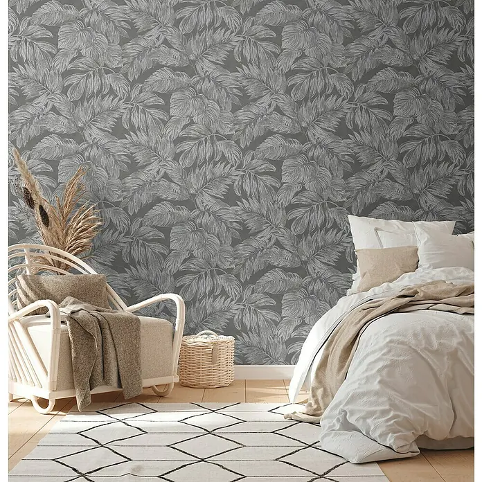 A.S. Création Casual Living Vliestapete - Anthrazit/Grau/Silber, Floral, 10,05 x 0,53 m - Nahaufnahme eines Schlafzimmers mit grauer Blättertapete, einem Sessel, einem Teppich und einem Bett mit weißer Bettwäsche.
