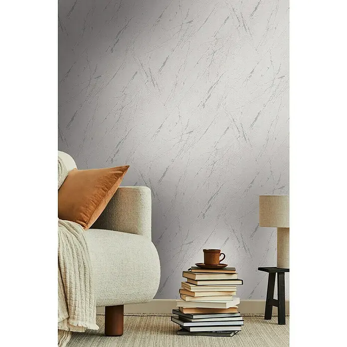 A.S. Création Casual Living Vliestapete Marmor - Silbergrau-Metallic, Steinoptik, 10,05 x 0,53 m - Nahaufnahme eines Raumes mit einer Couch mit Kissen, einem Bücherstapel mit Tasse und einer Lampe auf einem Beistelltisch.