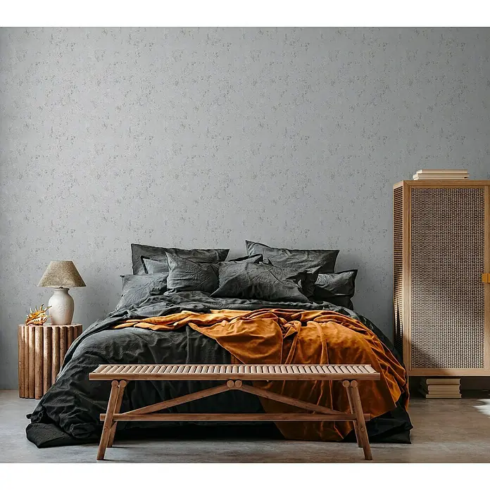 A.S. Création Casual Living Vliestapete Struktur - Grau/Silber/Metallic, Uni, 10,05 x 0,53 m - Ein gemütliches Schlafzimmer mit einem Doppelbett, grauer Bettwäsche und einer orangefarbenen Decke sowie einem Holzschrank.
