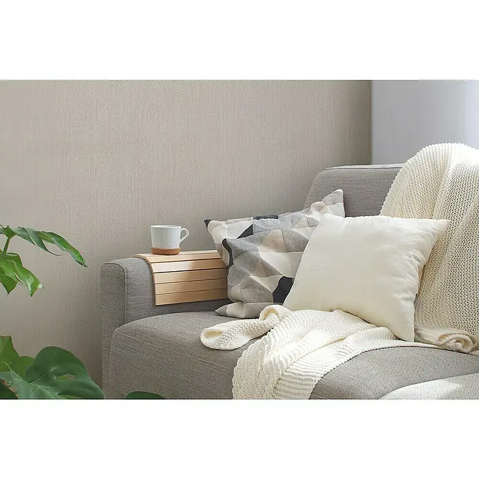 A.S. Création Casual Living Vliestapete Strukturtapete - Creme/Grau, Uni, 10,05 x 0,53 m - Ein graues Sofa mit Kissen, Decke und einem Beistelltisch mit einer Tasse darauf, für gemütliche Stunden im Wohnzimmer.