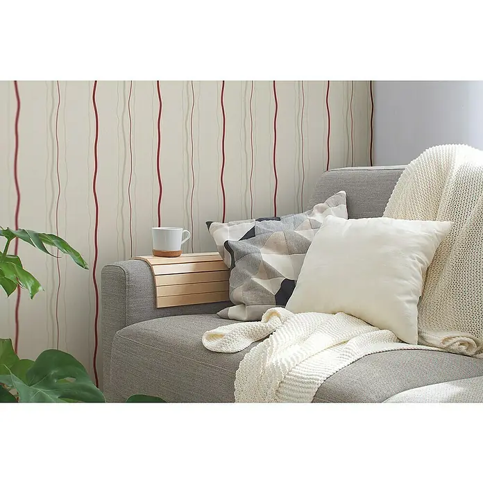 A.S. Création Casual Living Vliestapete - Creme/Rot/Beige, Streifen, 10,05 x 0,53 m - Nahaufnahme eines grauen Sofas mit Kissen, einer Strickdecke, einem hölzernen Armtablett und einer Tasse, vor einer Wand mit gestreiftem Tapetenmuster.