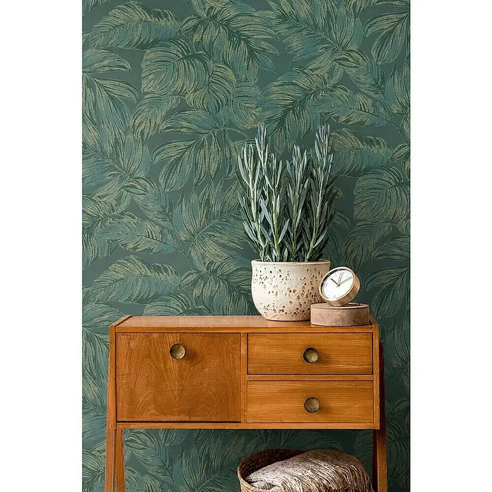 A.S. Création Casual Living Vliestapete - Grün/Gold, Floral, 10,05 x 0,53 m - Dekorative Kommode aus Holz mit Pflanze und Uhr darauf, vor einer grünen Blättertapete.