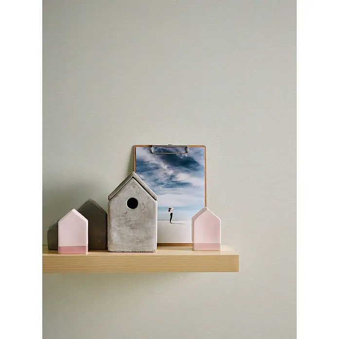 A.S. Création Casual Living Vliestapete - Creme/Weiß, Uni, 10,05 x 0,53 m - Dekorative Miniaturhäuser aus Beton und Holz auf einem hellen Holzregal, ergänzt durch ein gerahmtes Landschaftsbild.