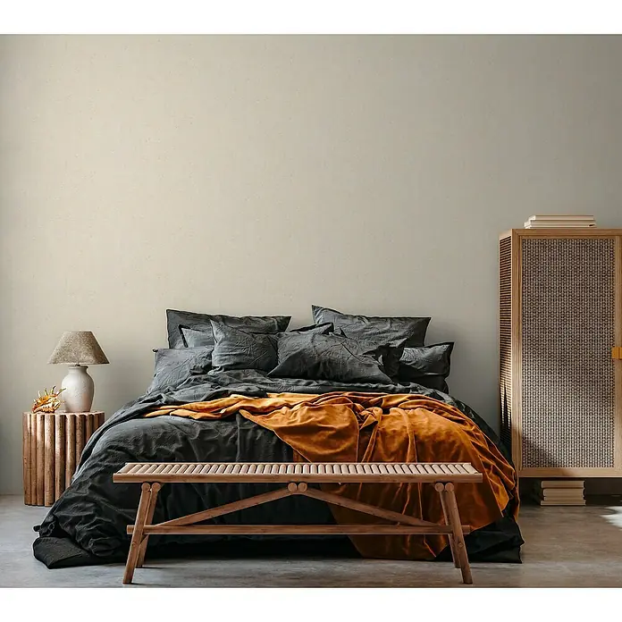 A.S. Création Casual Living Vliestapete Struktur - Beige, Uni, 10,05 x 0,53 m - Schlafzimmer-Interieur mit einem grauen Bett, einer orangefarbenen Decke und einem hölzernen Nachttisch mit Lampe.