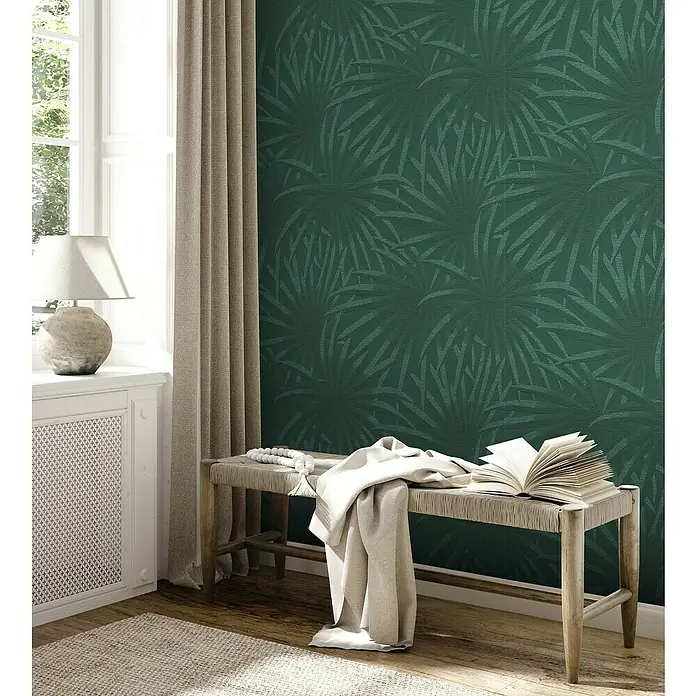 A.S. Création Casual Living Vliestapete Palmenblätter - Grün/Dunkelgrün, Floral, 10,05 x 0,53 m - Detailaufnahme eines Wohnraums mit grüner, floral gemusterter Tapete, einer Holzbank mit Überwurf und einem Fenster mit Vorhang.