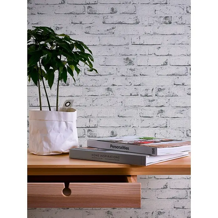 A.S. Création Casual Living Vliestapete New England Klinker - Grau/Anthrazit, Steinoptik, 10 x 0,53 m - Nahaufnahme eines hellen Holzschreibtischs mit Pflanze, Büchern und Schublade vor einer weißen Ziegelwand.