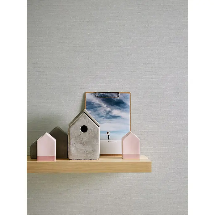 A.S. Création Casual Living Vliestapete - Strukturgrau, Uni, 10,05 x 0,53 m - Nahaufnahme von dekorativen Hausfiguren und einem Bild auf einem Holzregal vor einem strukturierten Hintergrund.