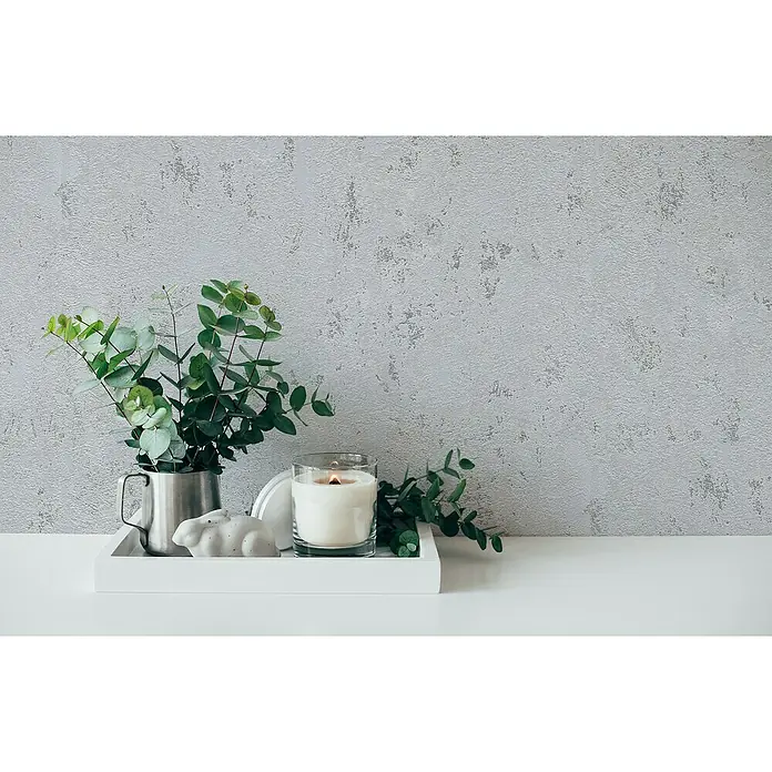 A.S. Création Casual Living Vliestapete Struktur - Grau/Silber/Metallic, Uni, 10,05 x 0,53 m - Stillleben mit Eukalyptus in einem Krug, einer Kerze und einer Hasenfigur auf einem weißen Tablett vor einer grauen Wand.