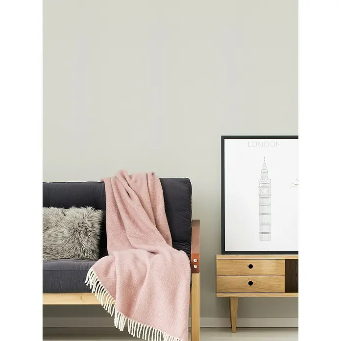 A.S. Création Casual Living Vliestapete - Creme-Weiß, Uni, 10,05 x 0,53 m - Gemütliches Wohnzimmer mit rosa Wolldecke auf einem grauen Sofa und einem Beistelltisch mit London-Poster.