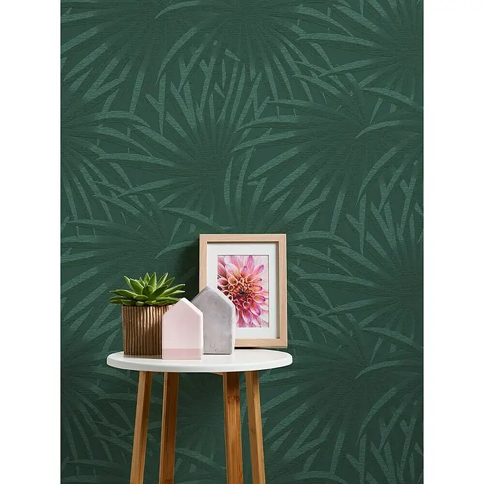 A.S. Création Casual Living Vliestapete Palmenblätter - Grün/Dunkelgrün, Floral, 10,05 x 0,53 m - Dekoratives Stillleben mit einer Zimmerpflanze, einem Bilderrahmen und Hausdekorationen auf einem kleinen Tisch vor einer grünen Blättertapete.