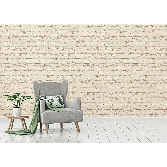 A.S. Création Casual Living Vliestapete New England Klinker - Weiß/Rot, Steinoptik, 10,05 x 0,53 m - Gemütliche Leseecke mit einem grauen Sessel, einem grünen Überwurf und einem kleinen Tisch mit einer Zimmerpflanze vor einer Backsteinwand.