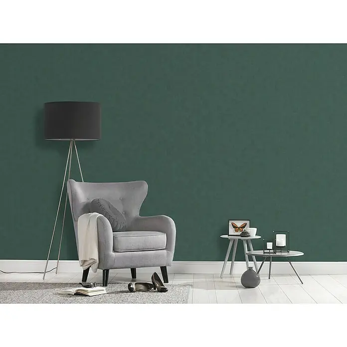 A.S. Création Casual Living Vliestapete - Grün, Uni, 10,05 x 0,53 m - Einrichtung mit einem stilvollen grauen Sessel mit Kissen und einer Stehlampe vor einer satten, grünen Wand.