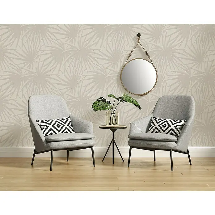 A.S. Création Casual Living Vliestapete Palmenblätter - Creme/Hellgrau, Floral, 10,05 x 0,53 m - Zwei graue Sessel mit geometrischen Kissen und einem Beistelltisch mit Pflanze vor einer Wand mit Palmenmuster.