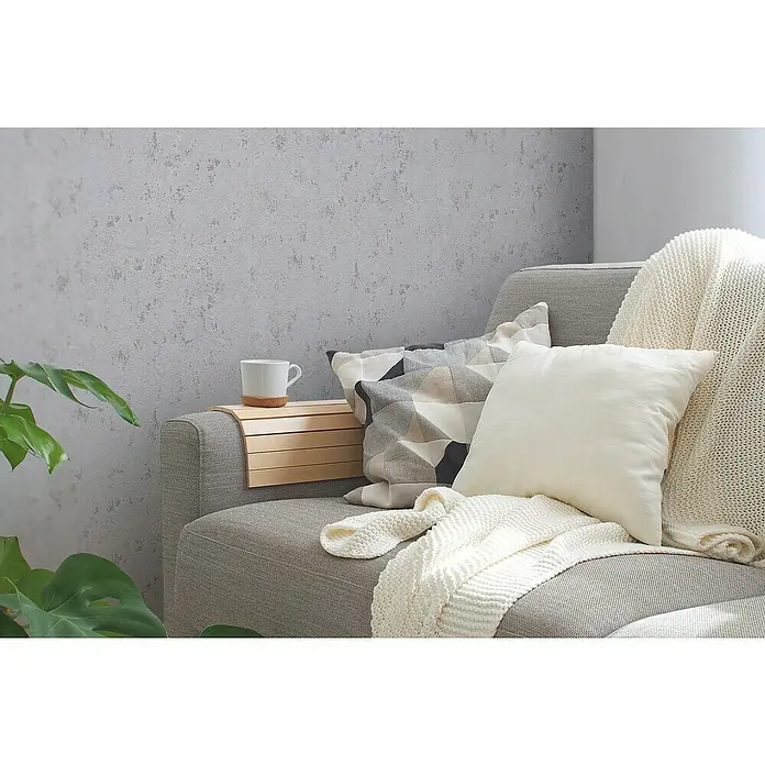 A.S. Création Casual Living Vliestapete Struktur - Grau/Silber/Metallic, Uni, 10,05 x 0,53 m - Bequemes graues Sofa mit hellen Kissen und einer Strickdecke, ergänzt durch eine Holzablage mit einer weißen Tasse.