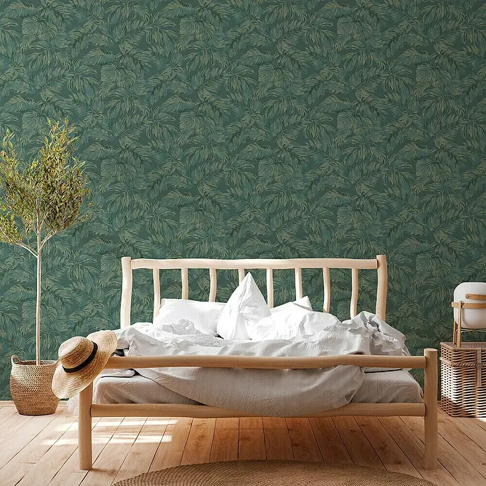 A.S. Création Casual Living Vliestapete - Grün/Gold, Floral, 10,05 x 0,53 m - Schlafzimmer mit einem Holzbett unter grüner Blatt-Tapete. Ein Strohhut liegt auf dem Bett und eine Topfpflanze steht neben dem Bett.
